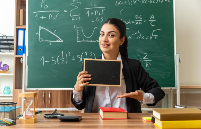 smiling young female teacher sits at table with school tools holding and points at mini blackboard in classroom